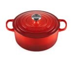 A red Le Creuset cast iron Dutch oven with a lid and two side handles, featuring a silver knob on top, shown against a white background.