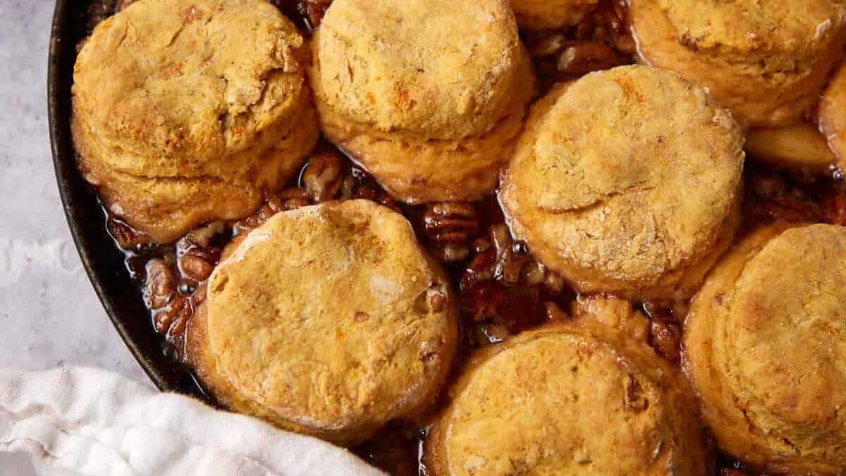 A cast iron skillet filled with golden-brown sweet potato biscuits baked atop a gooey apple cobbler filling, with a beige striped kitchen towel partially visible on the left side.