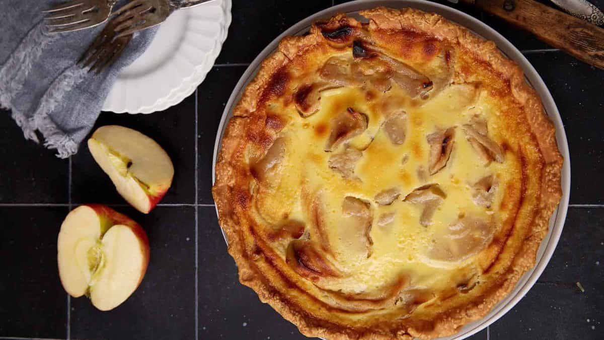 A baked apple custard tart in a round pie dish sits on a dark tiled surface, with a sliced apple and a stack of plates with forks nearby.