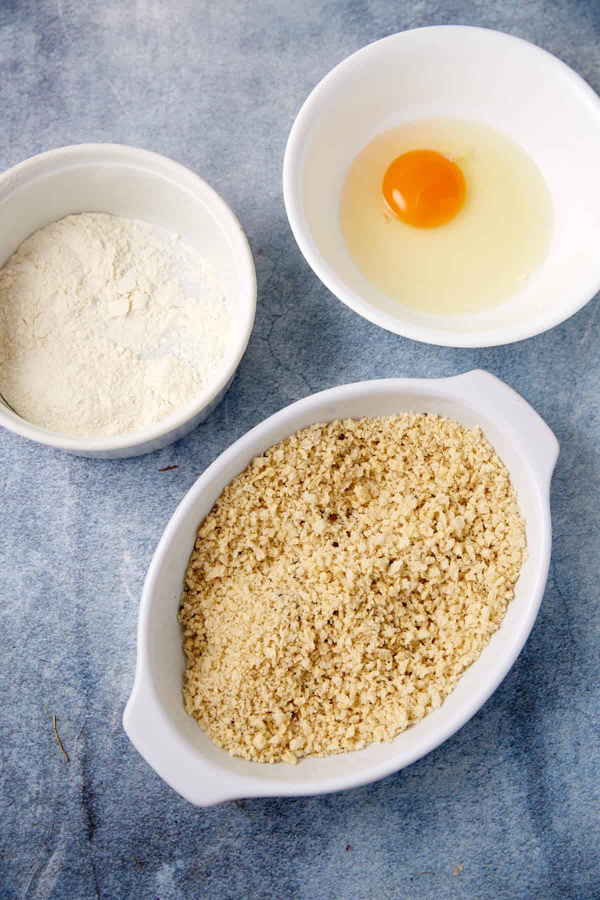 Three white bowls on a blue surface: one contains an egg with yolk intact, another has flour, and the third is filled with light tan breadcrumbs.