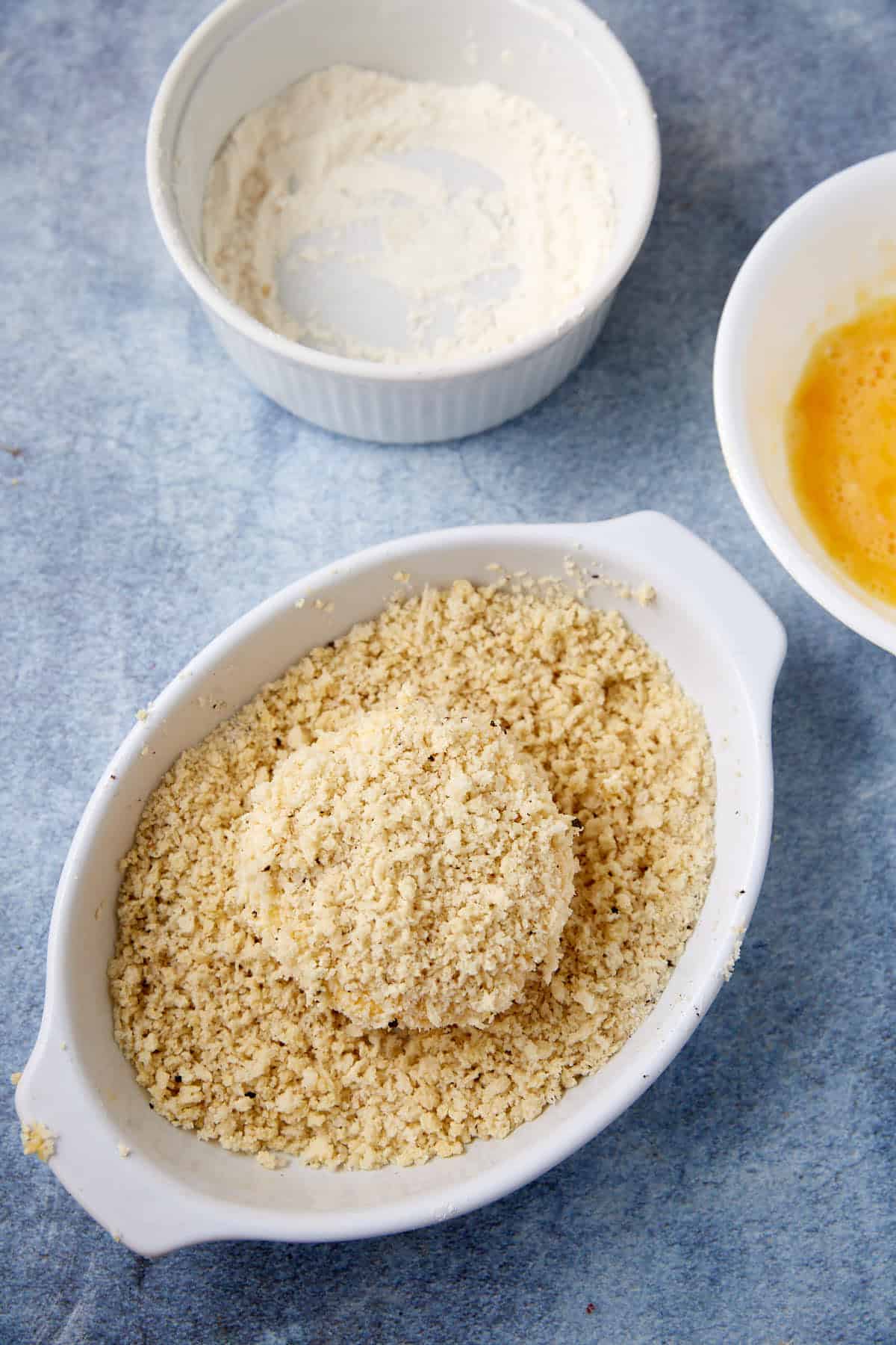 A breaded food item sits in a white dish filled with breadcrumbs. Nearby, a bowl of beaten eggs and a dish of flour suggest a breading station on a blue surface.