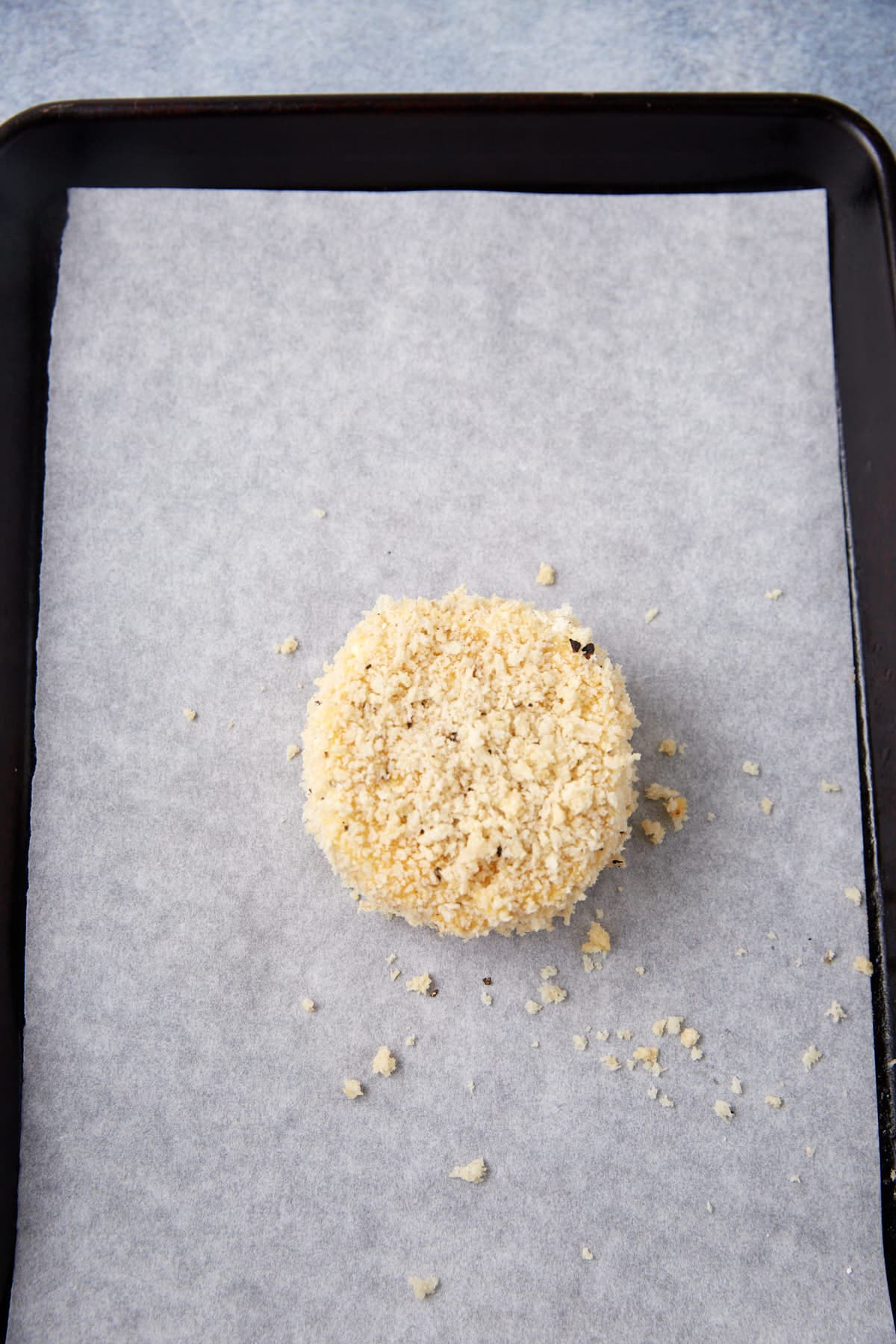 A single round, breaded item sits on a parchment-lined baking tray with some crumbs scattered around it.
