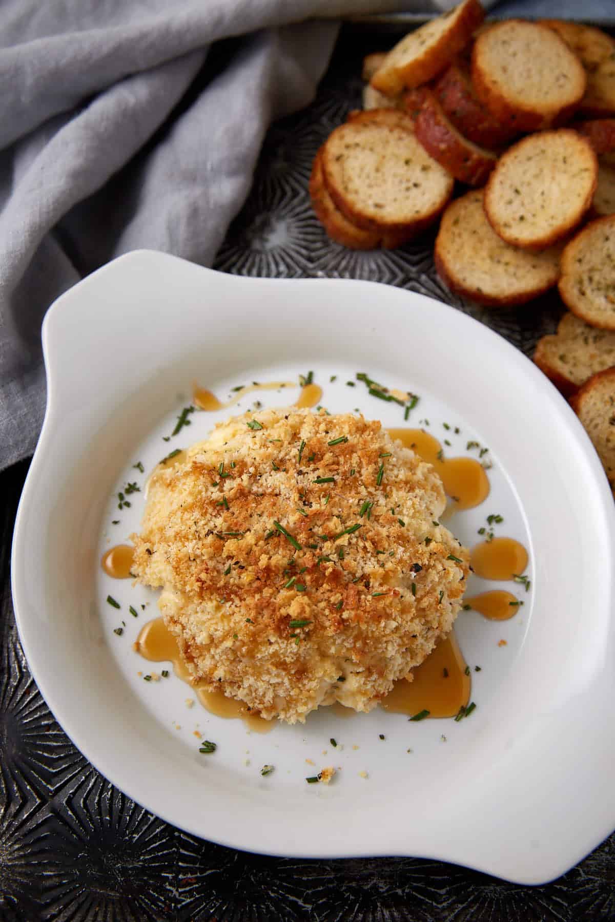 A round, baked cheese dip topped with golden breadcrumbs and herbs sits in a white dish, drizzled with honey. Slices of toasted baguette are arranged nearby on a textured dark surface.