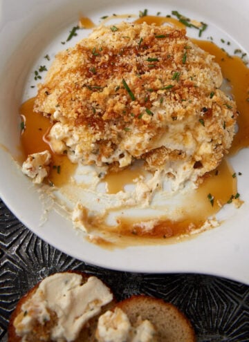 A baked crab dip with a golden breadcrumb topping sits in a white dish, garnished with herbs and surrounded by a light sauce. A piece of bread with dip spread on it is visible in the foreground.