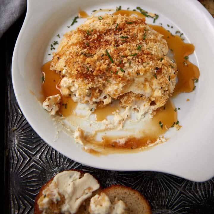 A baked crab dip with a golden breadcrumb topping sits in a white dish, garnished with herbs and surrounded by a light sauce. A piece of bread with dip spread on it is visible in the foreground.