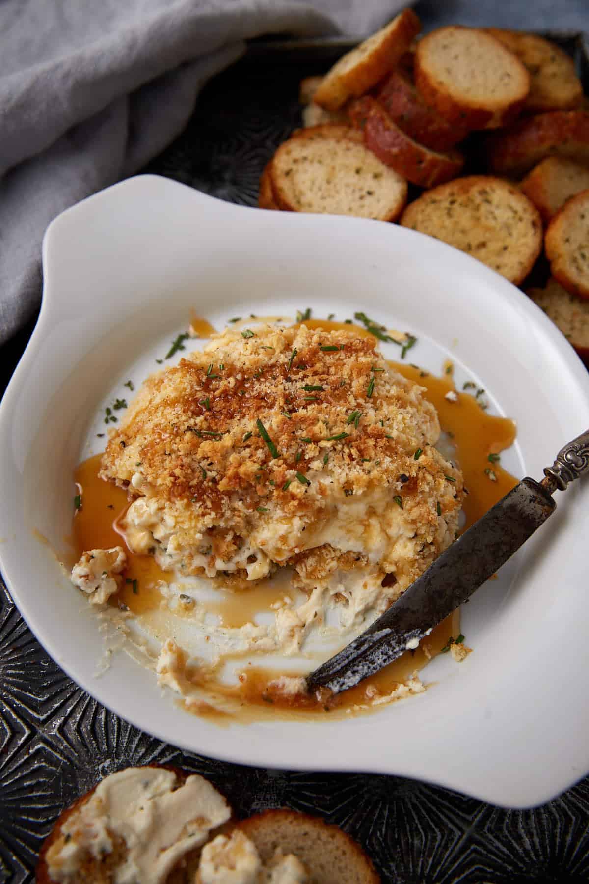 A partially eaten baked cheese dip topped with golden breadcrumbs in a white dish, with a spreading knife and sliced toasted baguette rounds on the side.
