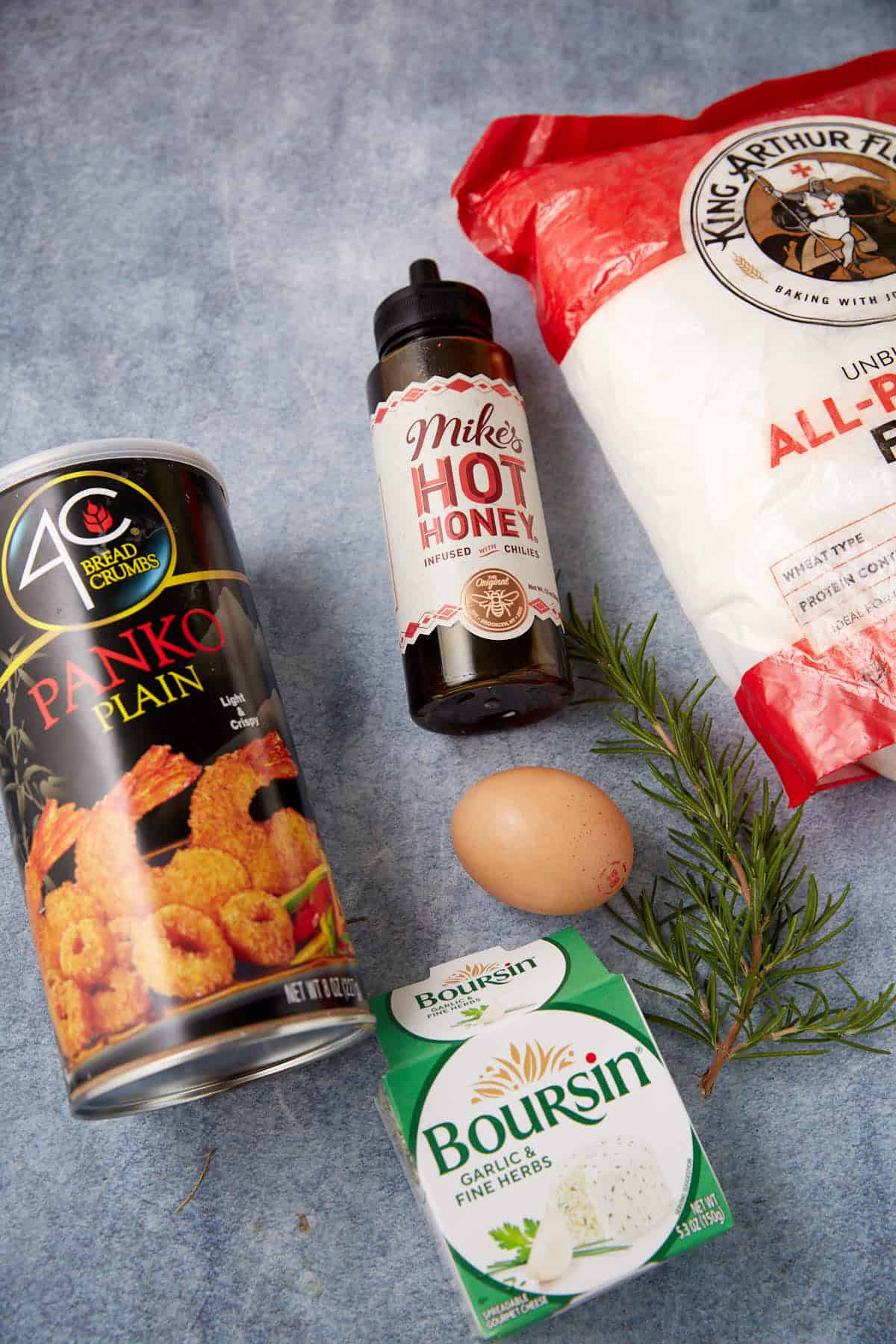 A can of panko breadcrumbs, a bottle of hot honey, a bag of all-purpose flour, a fresh egg, a sprig of rosemary, and a box of Boursin garlic & herb cheese are arranged on a blue surface.