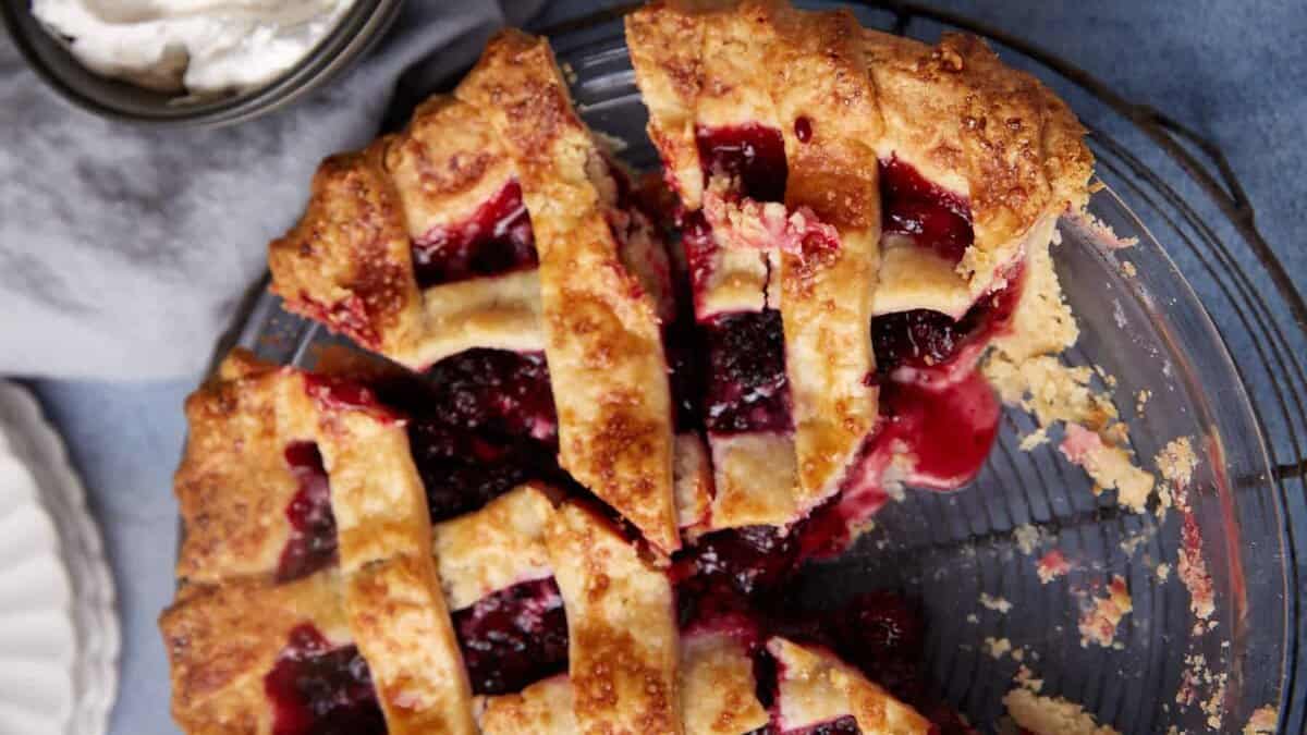 A top-down view of a partially eaten blackberry pie with a lattice crust on a wire cooling rack. The pie has a crispy golden-brown crust with juicy, dark purple filling. A bowl of whipped cream is placed nearby. Crumbs and a piece of pie are visible on the table.