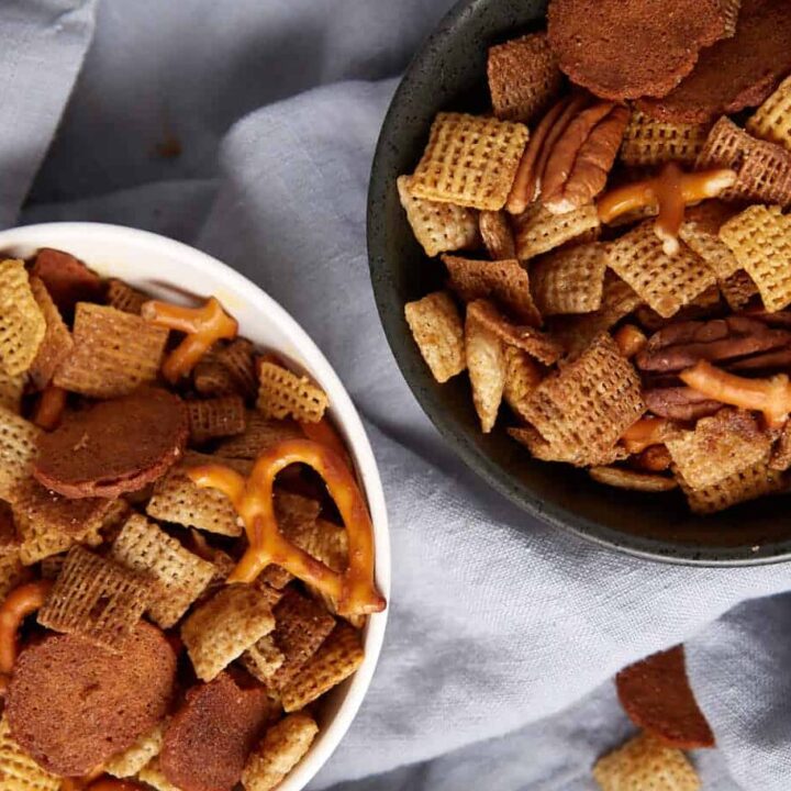 Three bowls filled with a bold Chex Mix recipe—Chex cereal, pretzels, rye chips, and nuts—sit on a light gray cloth, with some pieces scattered around the bowls.