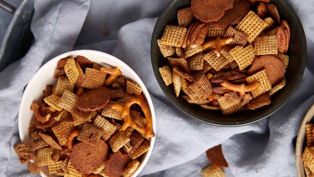 Three bowls filled with a bold Chex Mix recipe—Chex cereal, pretzels, rye chips, and nuts—sit on a light gray cloth, with some pieces scattered around the bowls.