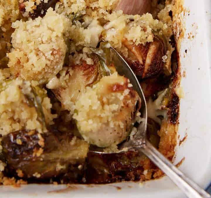 A close-up of a baked casserole dish filled with Brussels sprouts topped with golden, crispy breadcrumbs. A serving spoon is partly inserted, showing the creamy interior.