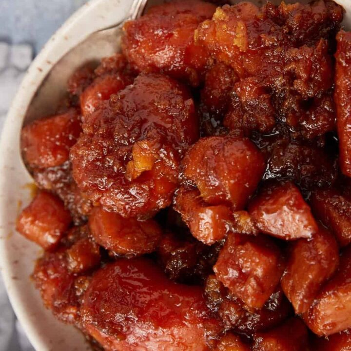 A bowl filled with southern candied yams and sweet potatoes in a thick, caramelized sauce, sitting on a light blue surface with a gray cloth in the background.