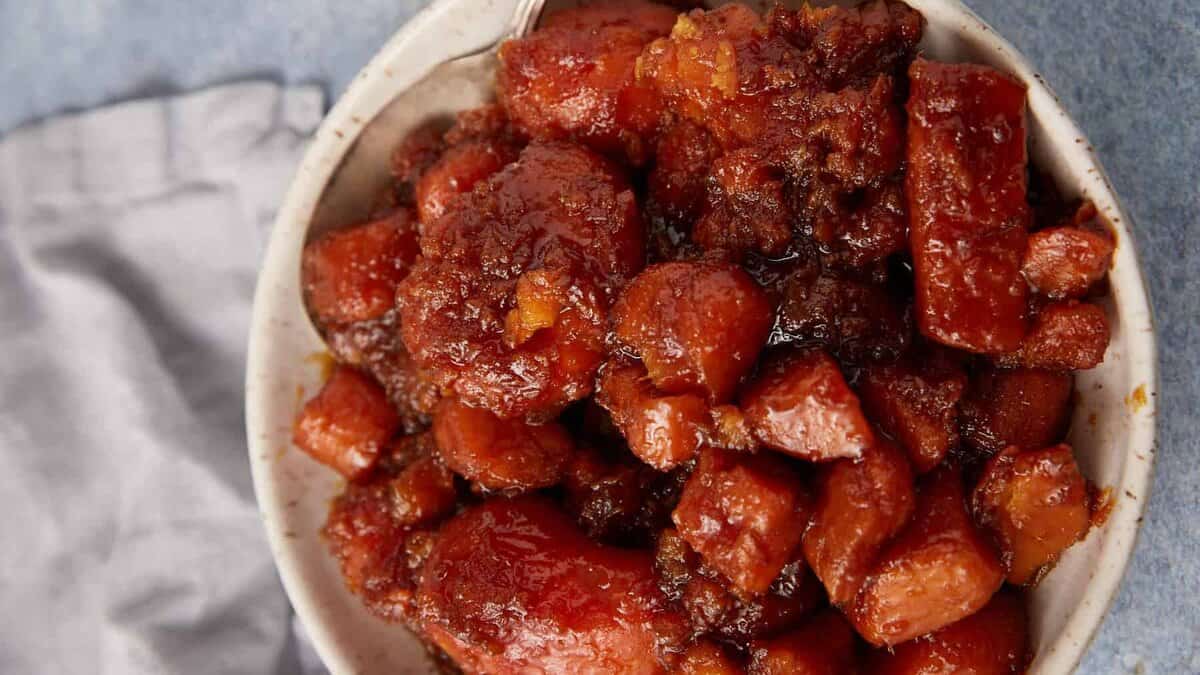 A bowl filled with southern candied yams and sweet potatoes in a thick, caramelized sauce, sitting on a light blue surface with a gray cloth in the background.