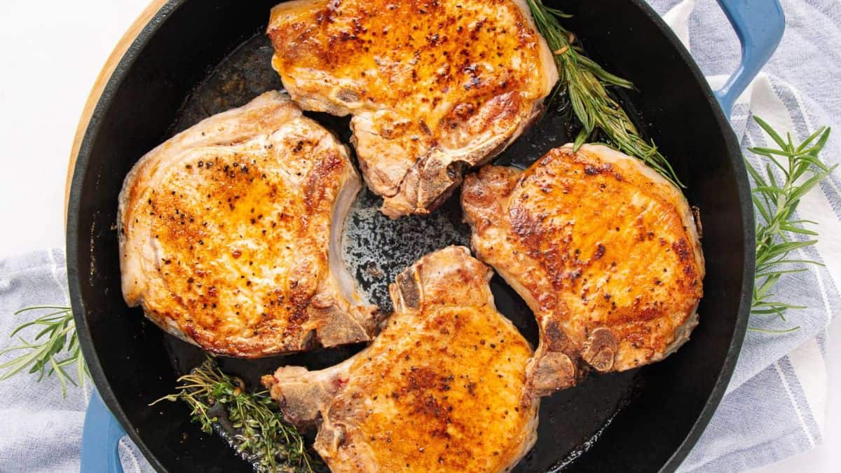 Four golden-brown, seared pork chops in a black skillet, garnished with fresh rosemary and thyme, make for irresistible turkey-free mains, all resting on a light gray cloth.
