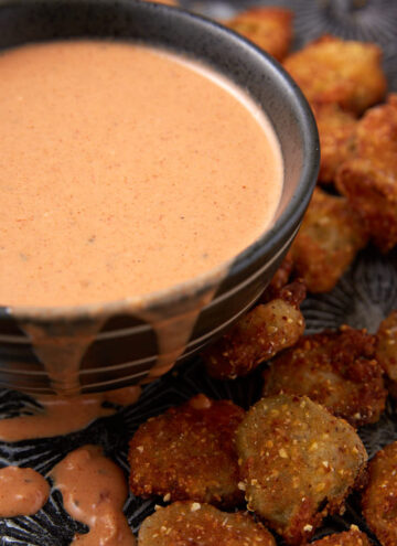 A black bowl filled with creamy, pinkish dipping sauce, with some sauce dripping down the side, sits next to a pile of crispy, golden-brown fried pickles on a dark textured surface.