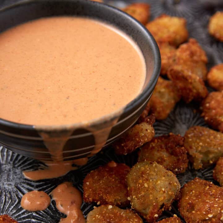 A black bowl filled with creamy, pinkish dipping sauce, with some sauce dripping down the side, sits next to a pile of crispy, golden-brown fried pickles on a dark textured surface.