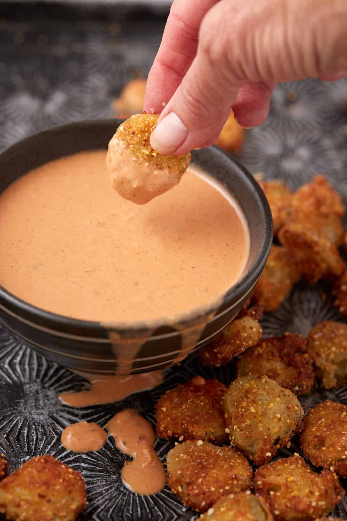 A hand dips a crispy fried pickle slice into a bowl of creamy, orange-colored sauce, with more fried pickles and sauce drips on a decorative black tray.