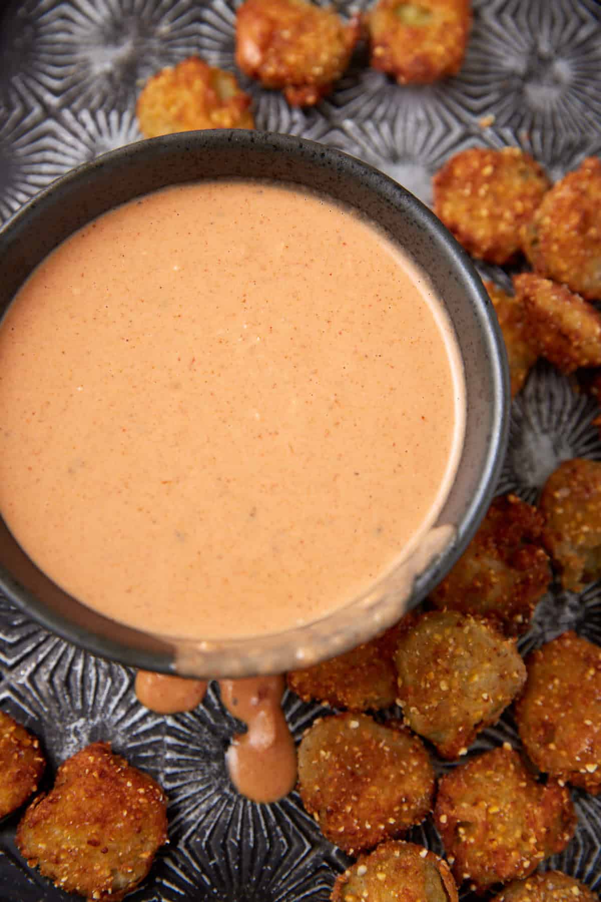 A black bowl filled with creamy, orange dipping sauce sits on a tray surrounded by fried pickle chips; some sauce drips down the side of the bowl.