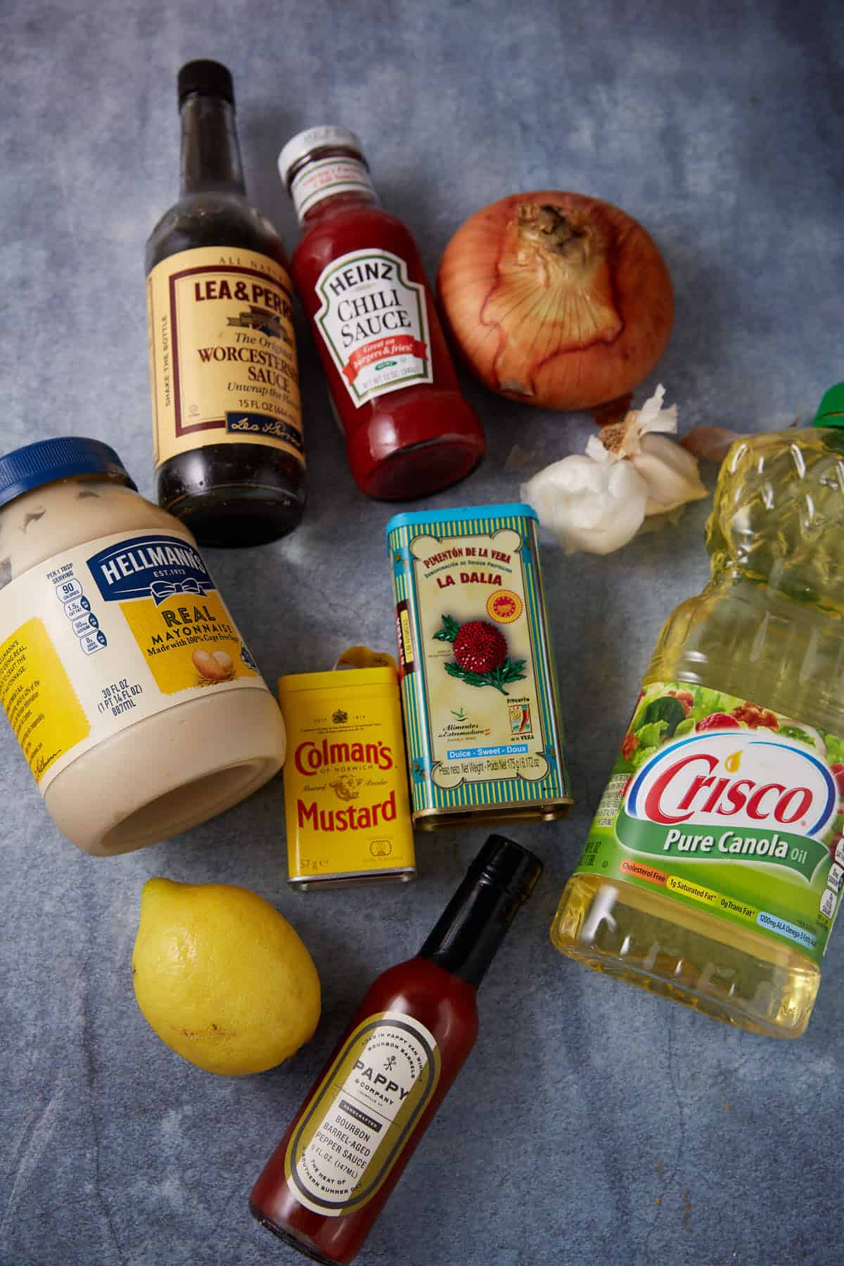 Assorted ingredients on a gray surface, including mayonnaise, Worcestershire sauce, chili sauce, an onion, garlic, canola oil, a lemon, hot sauce, powdered mustard, and a tin of Spanish paprika.