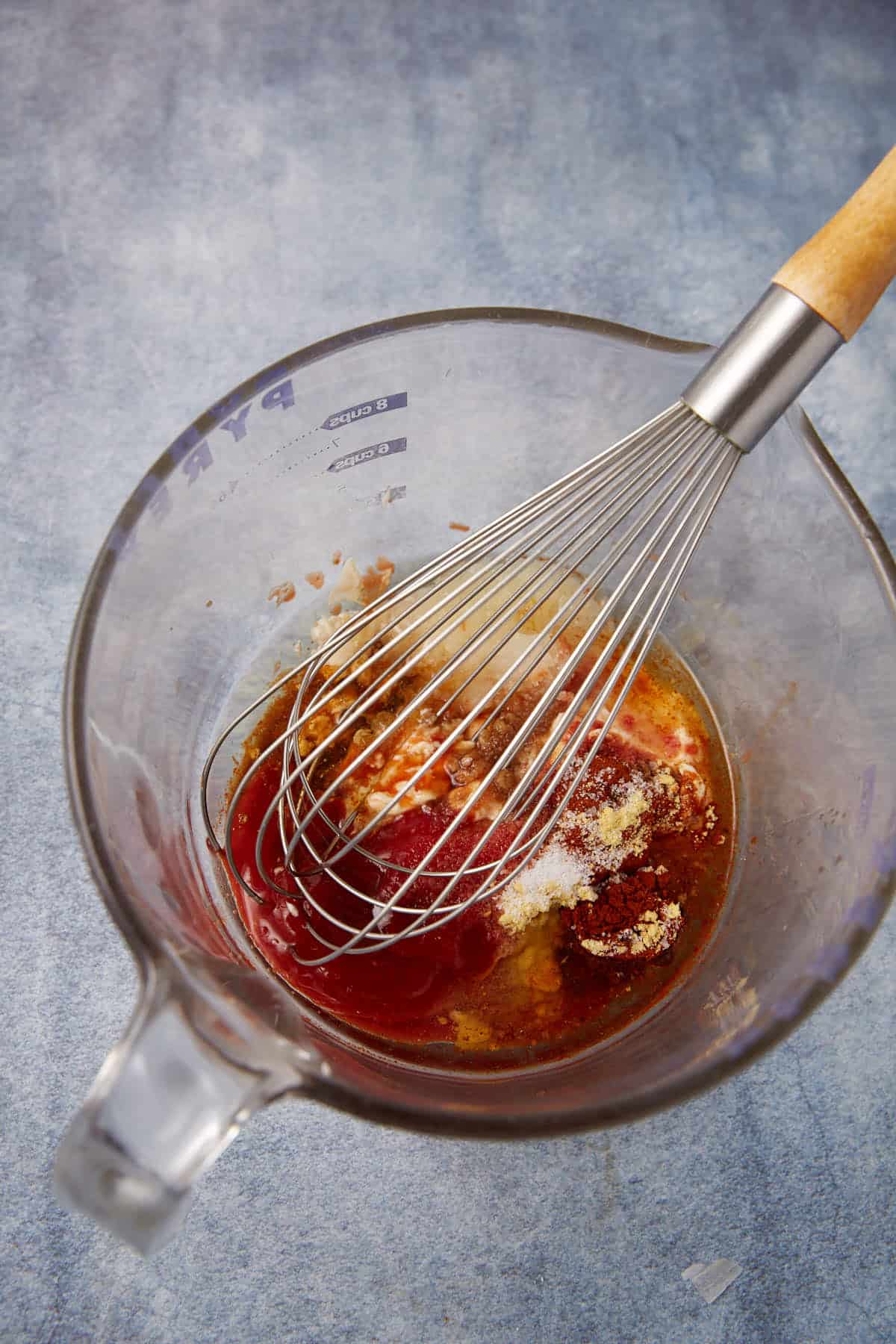 A glass mixing bowl with a whisk and various reddish and white ingredients, including spices and liquid, ready to be mixed, sits on a blue-gray countertop.