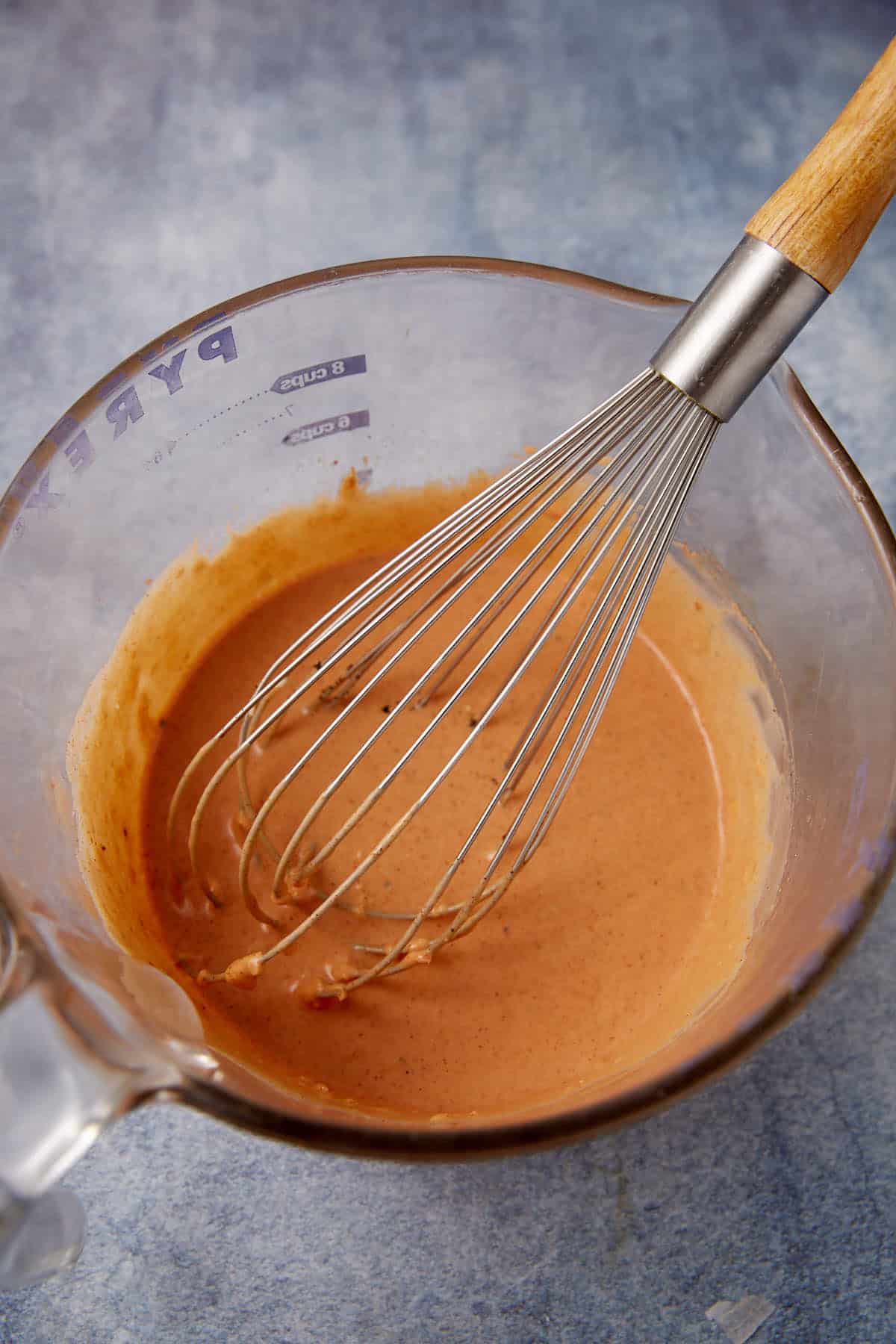 A metal whisk with a wooden handle rests in a glass measuring cup containing a smooth, light brown sauce or batter on a blue surface.