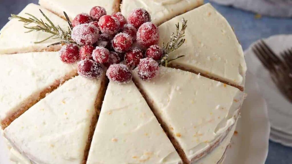 A round, frosted cake sliced into pieces, topped with sugared cranberries and rosemary sprigs, is displayed on a white plate with forks in the background—one of those holiday cakes that steal the show on dessert tables.