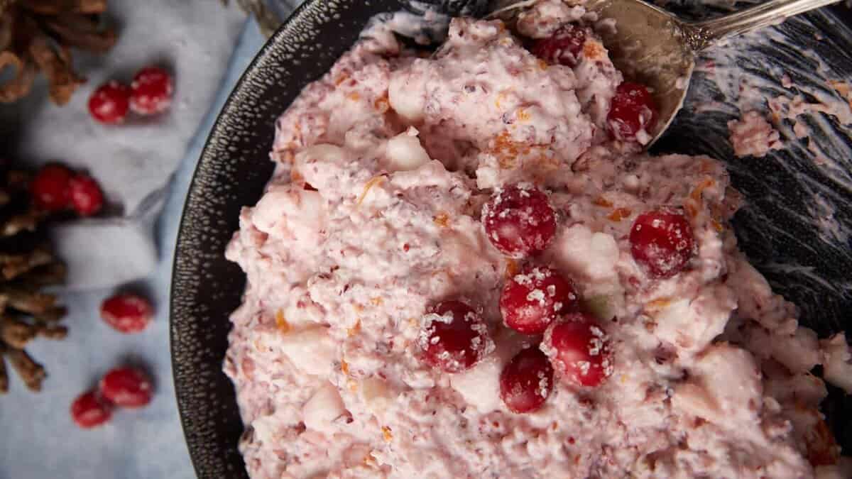 A close-up of cranberry fluff salad in a black dish, garnished with whole cranberries. A silver spoon is resting in the mixture, and pinecones are visible in the background.