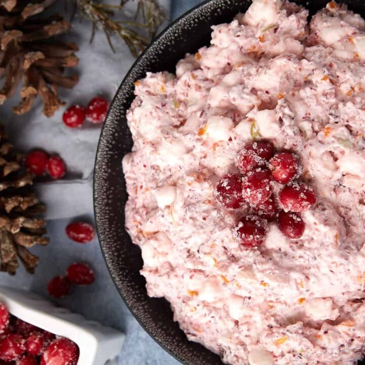 A black bowl filled with pink cranberry fluff salad topped with sugared cranberries, next to a white dish of cranberries and two pinecones on a blue-gray surface.
