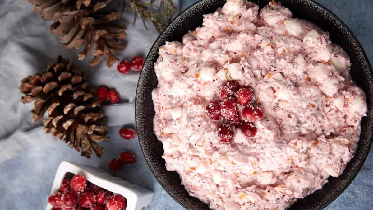 A black bowl filled with pink cranberry fluff salad topped with sugared cranberries, next to a white dish of cranberries and two pinecones on a blue-gray surface.