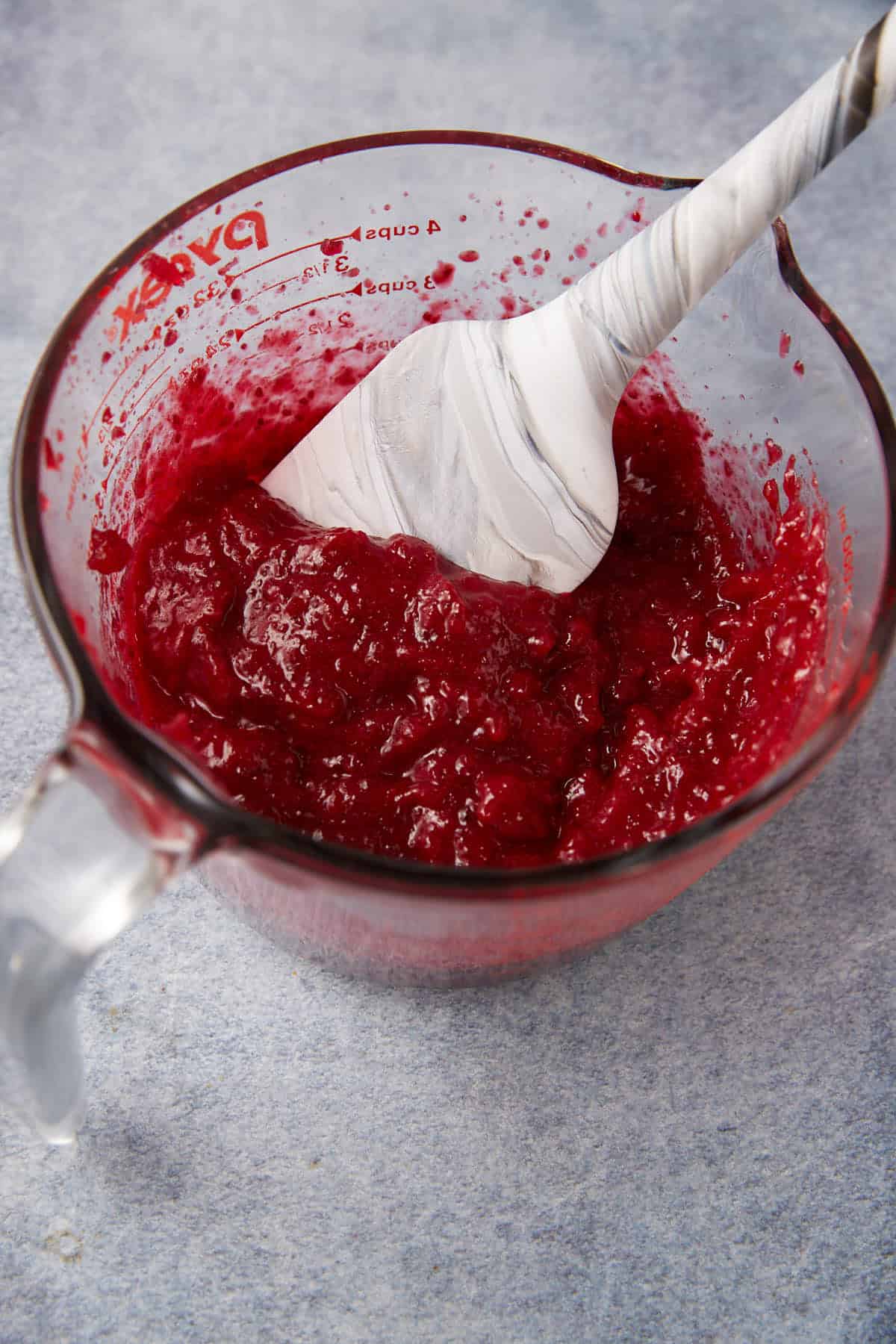 A glass measuring cup filled with thick red cranberry sauce is being stirred with a white and gray marbled spatula, sitting on a light blue surface.