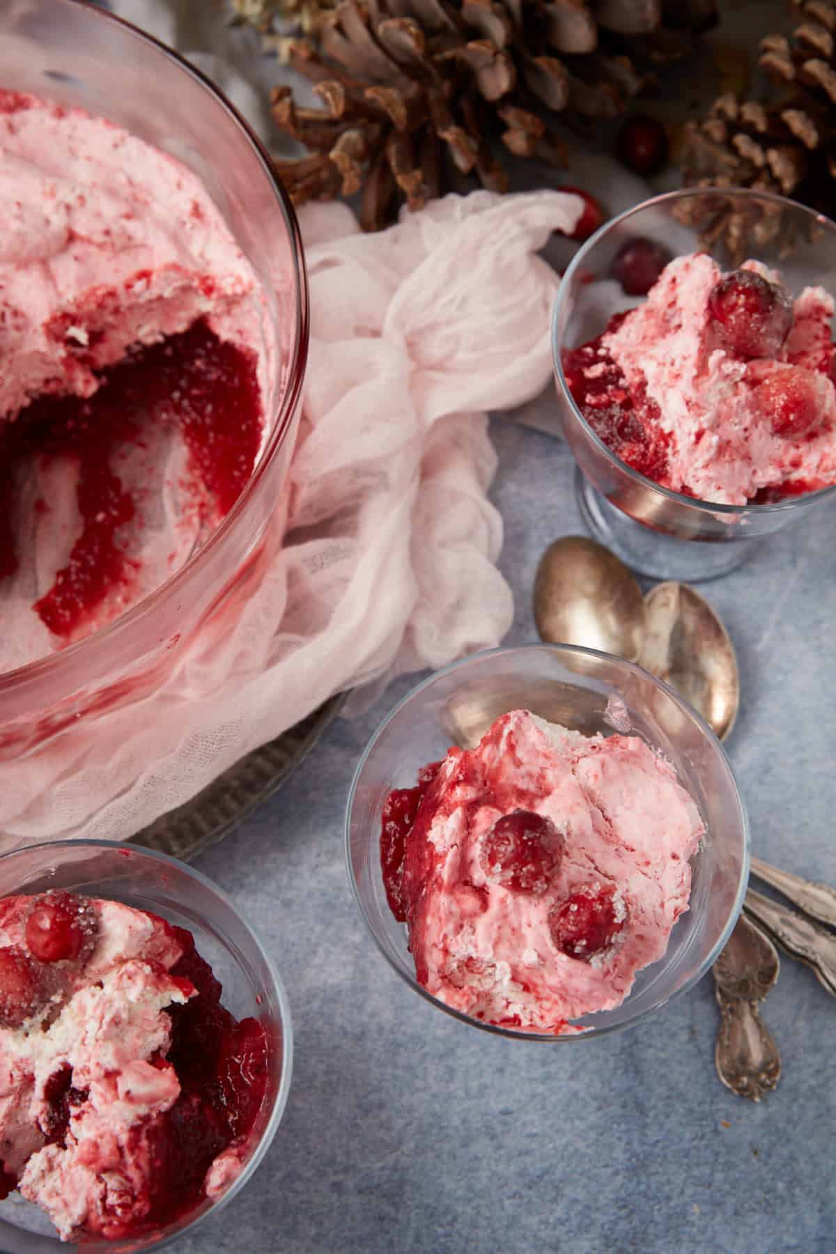 Three glass cups and a large bowl filled with a pink, creamy cranberry dessert sit on a blue surface, surrounded by vintage silver spoons, pinecones, and a light pink cloth.