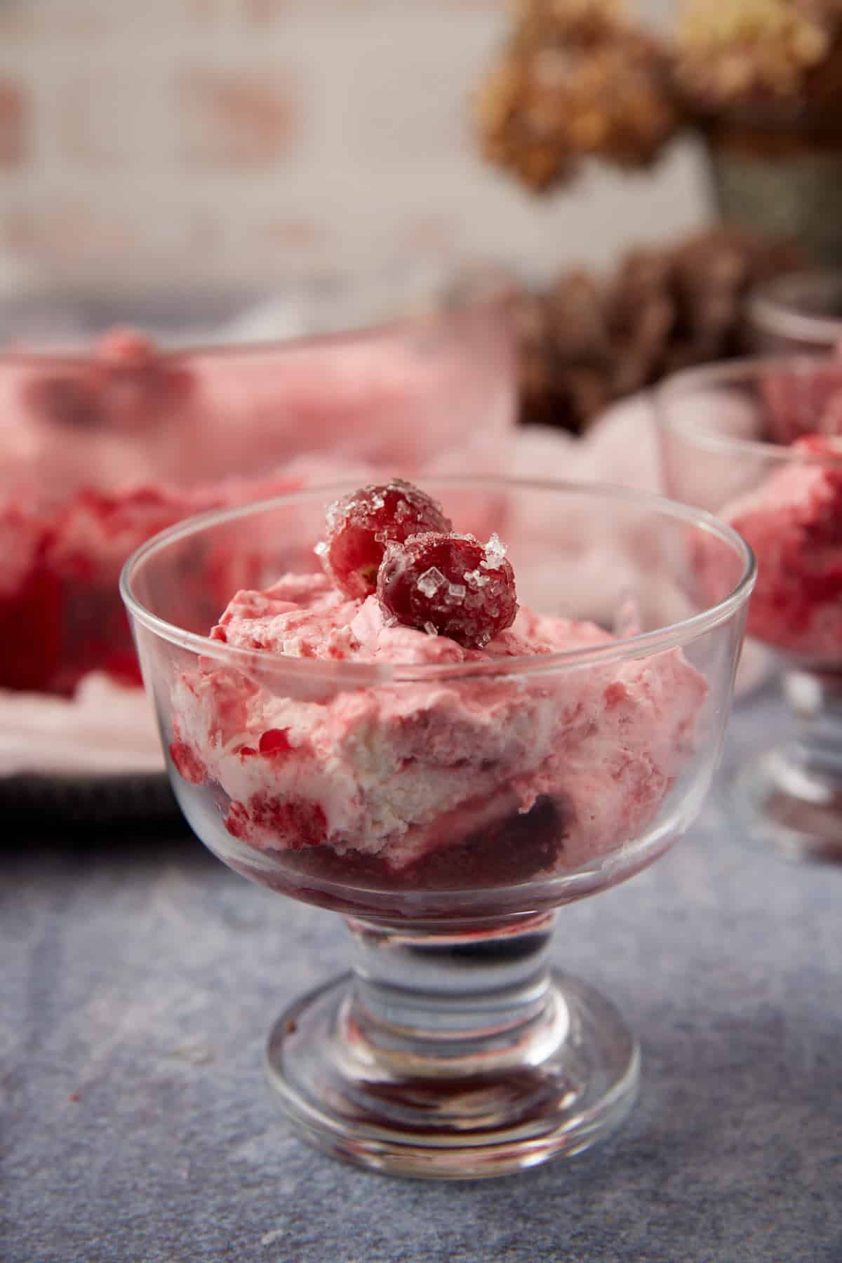 A glass dessert dish filled with a creamy pink dessert topped with sugared cranberries, with more servings and blurred background elements visible behind it.