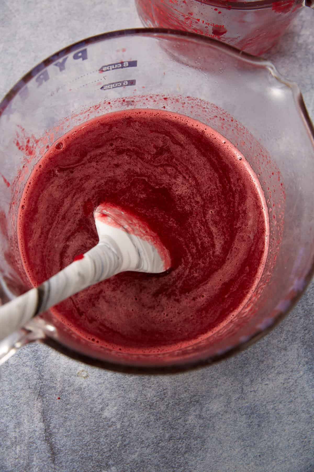 A glass measuring cup filled with a deep red liquid being stirred with a white marbled spoon on a gray surface.