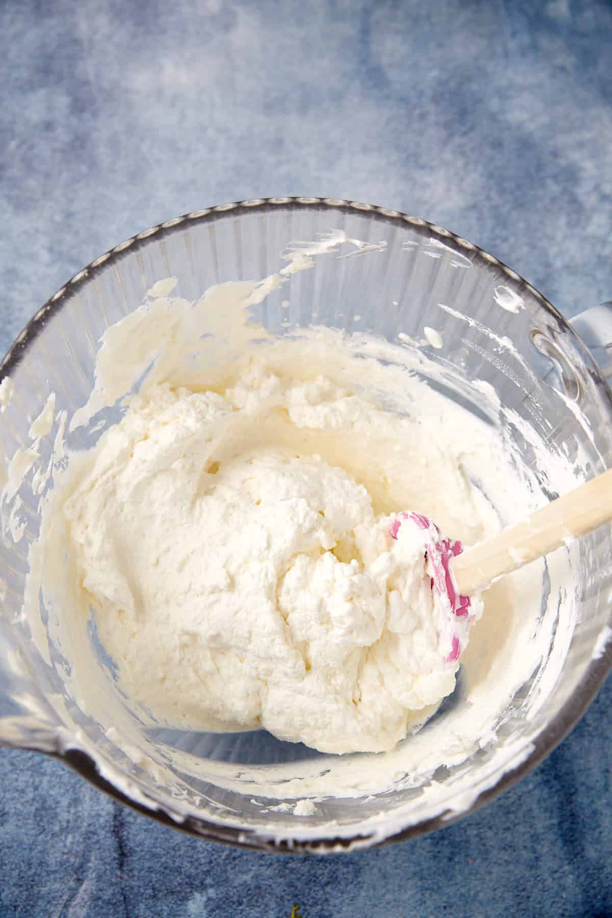 A glass mixing bowl containing a thick, creamy mixture is set on a blue surface. A spatula with a white and pink tip rests inside the bowl, partially covered in the mixture.