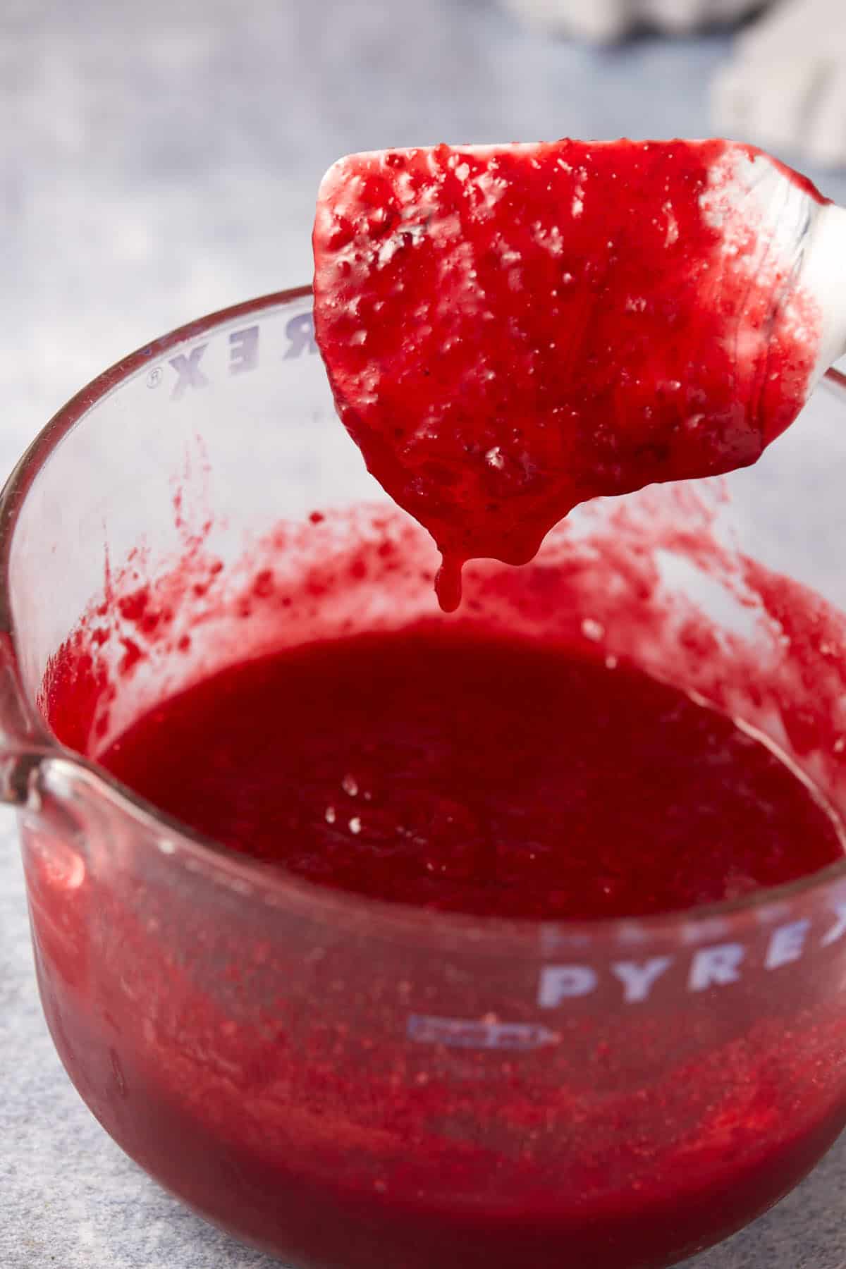 A spatula holds up a thick, red raspberry sauce over a glass Pyrex measuring cup filled with more of the chunky sauce. The background is out of focus.