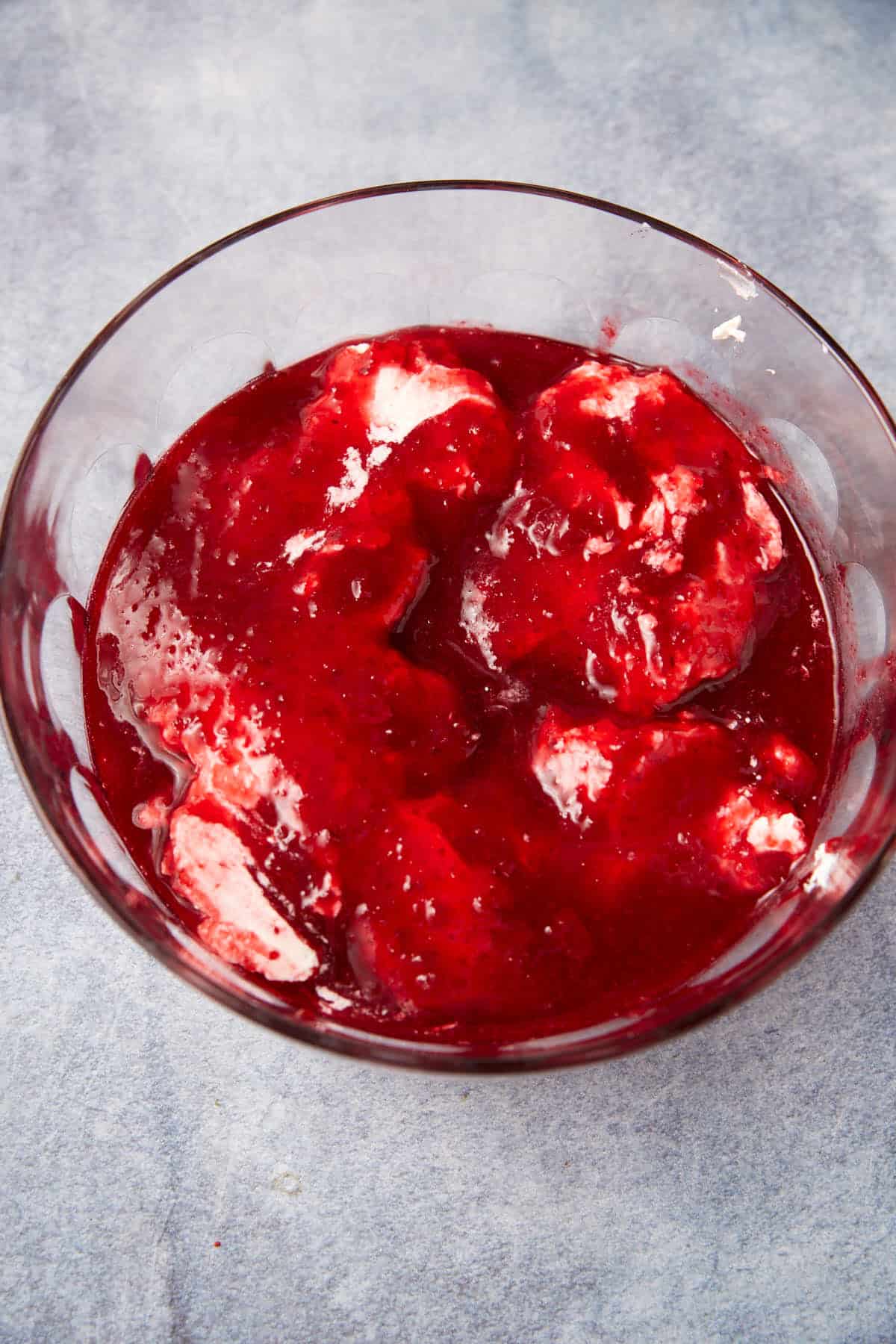 A glass bowl filled with red berry sauce poured over scoops of white ice cream, set on a light grey surface.