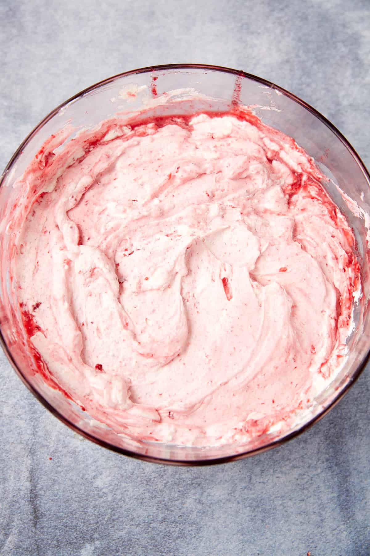 A glass bowl filled with a pink, fluffy mixture that appears to be whipped cream blended with crushed strawberries, sitting on a light gray surface.