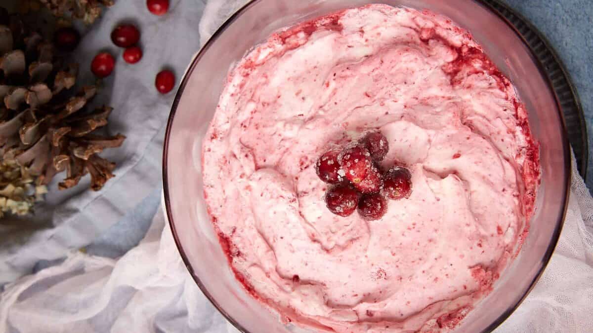 A glass bowl filled with pink cranberry fluffy salad, topped with sugared cranberries, sits on a white cloth with pine cones and loose cranberries nearby.