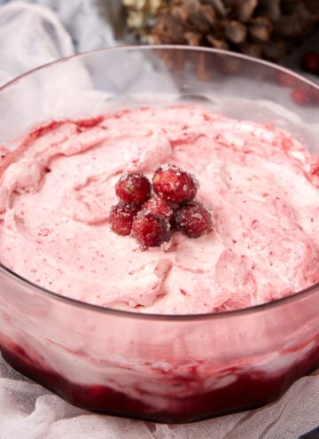A clear glass bowl filled with pink, swirled cranberry fluff topped with sugared cranberries, set on a light cloth with pinecones in the background.