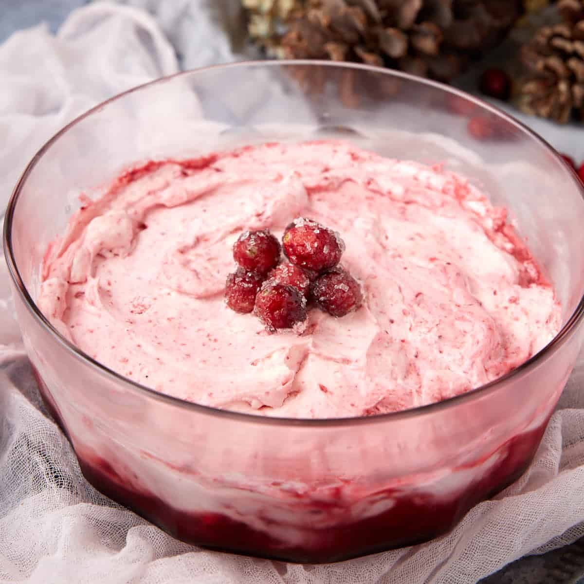 A clear glass bowl filled with pink, swirled cranberry fluff topped with sugared cranberries, set on a light cloth with pinecones in the background.