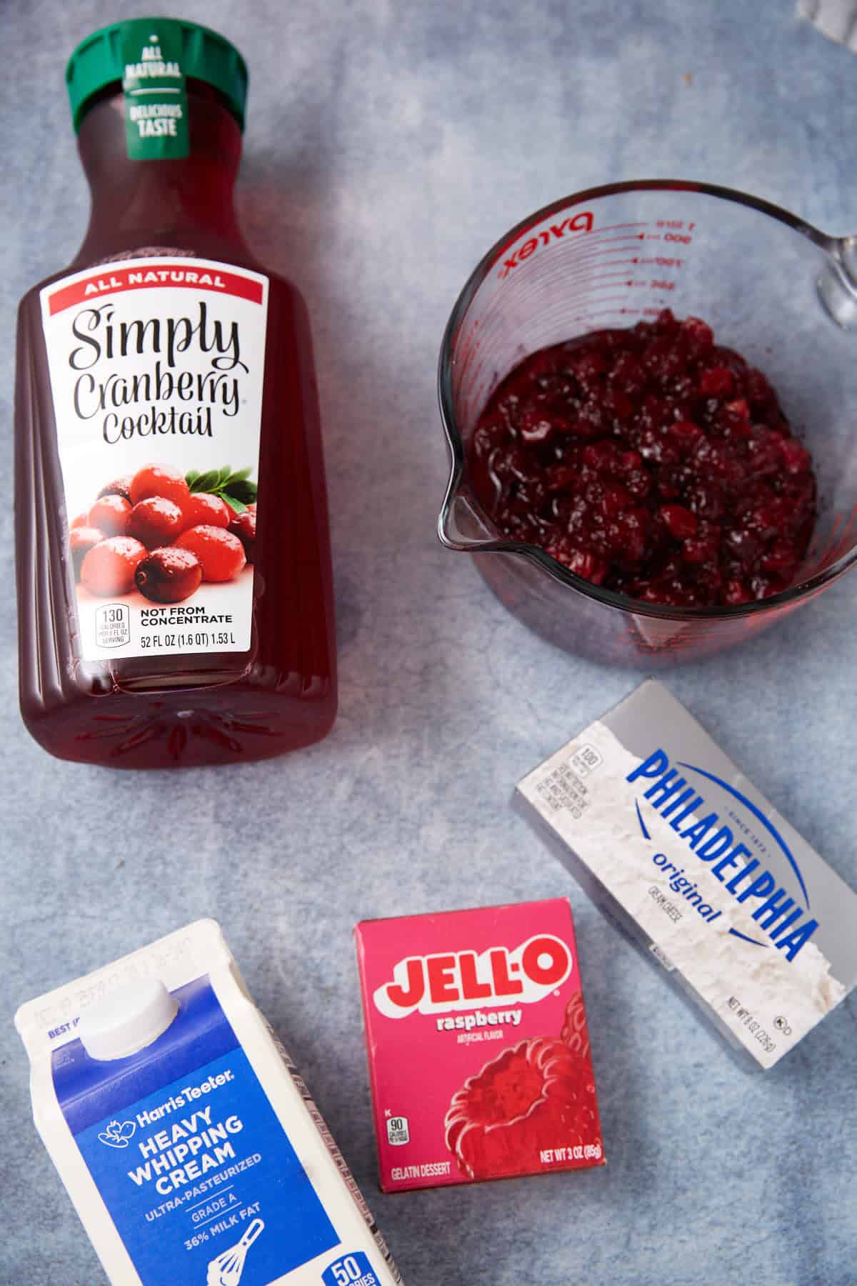 A bottle of cranberry cocktail, a measuring cup with cranberry sauce, a block of cream cheese, a carton of heavy whipping cream, and a box of raspberry Jell-O on a light blue surface.