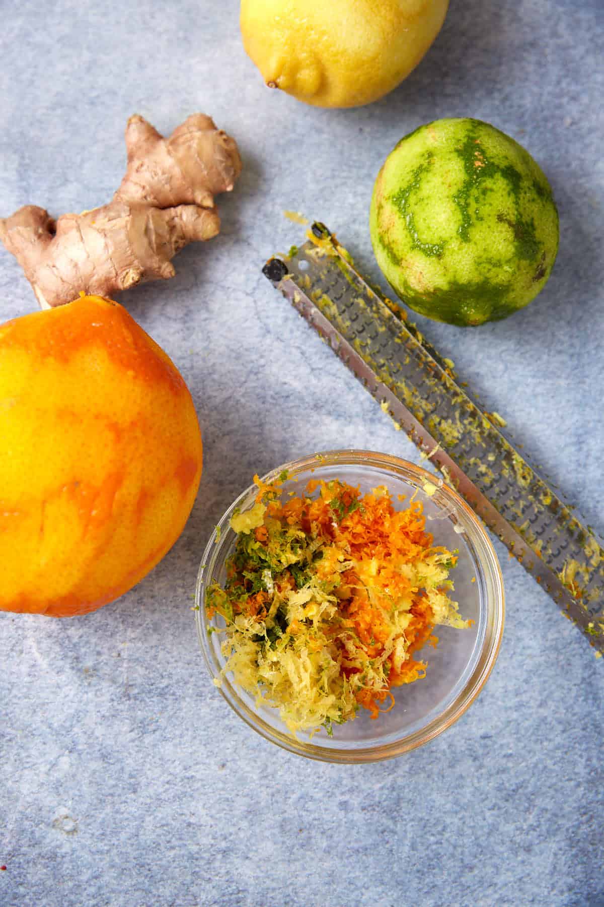 A bowl of mixed citrus zest sits beside a zester, with whole ginger, a lemon, a lime, and an orange on a gray surface.