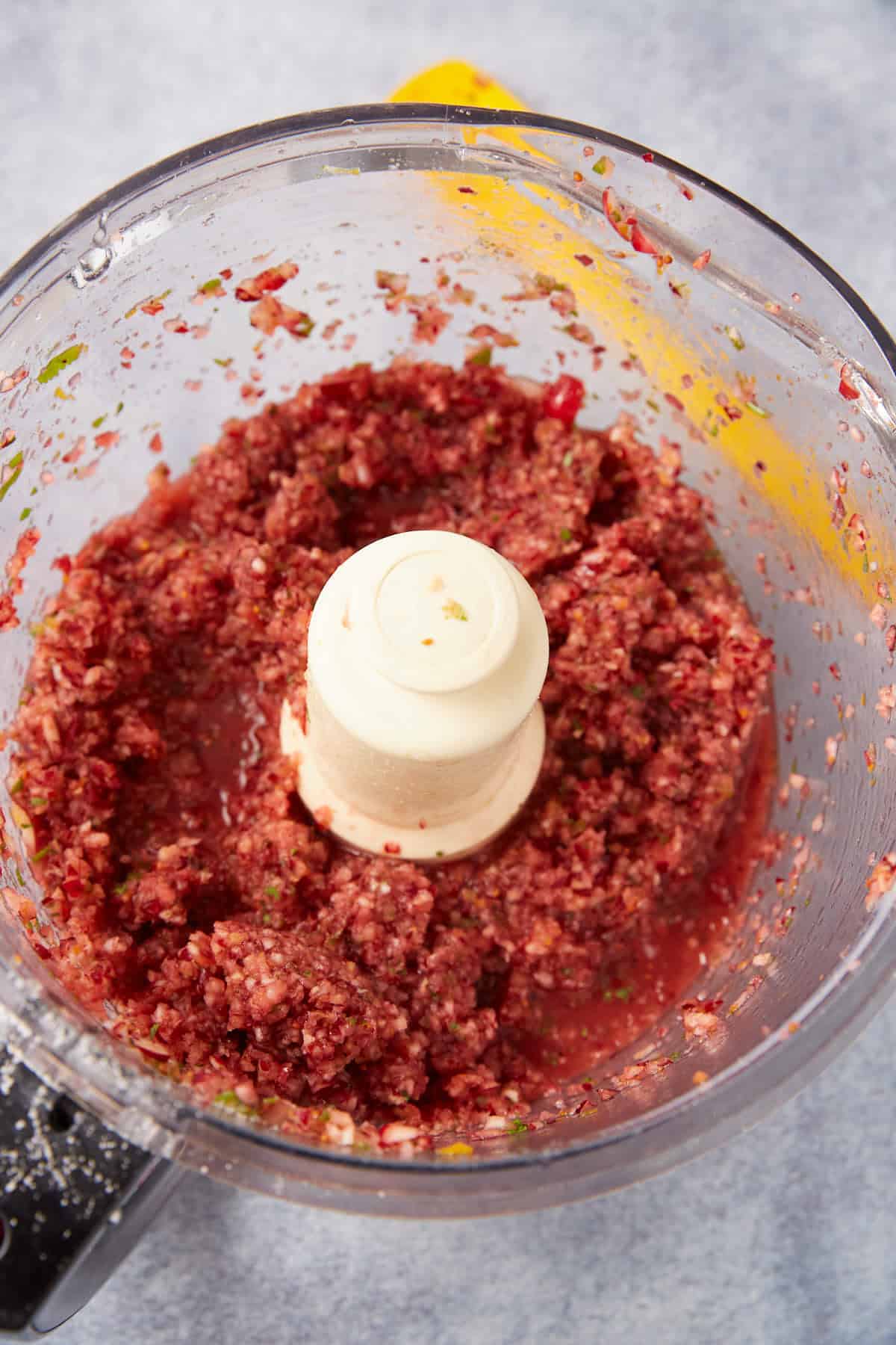 A food processor bowl containing finely chopped red and green ingredients, possibly a fresh salsa or relish, with visible bits and juices around the blade.