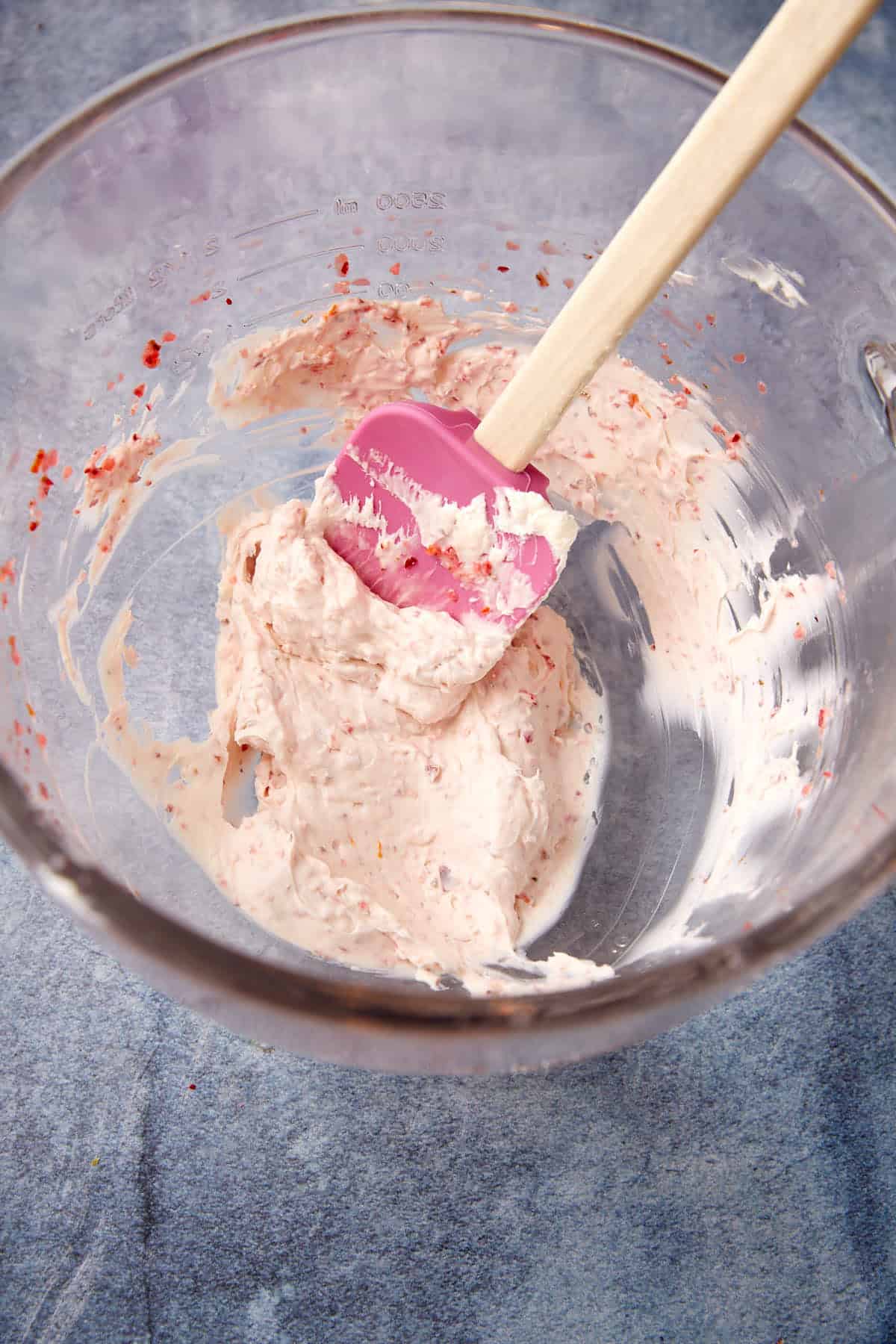 A glass mixing bowl containing a light pink creamy mixture with red specks, being stirred by a pink spatula with a wooden handle, set on a blue surface.