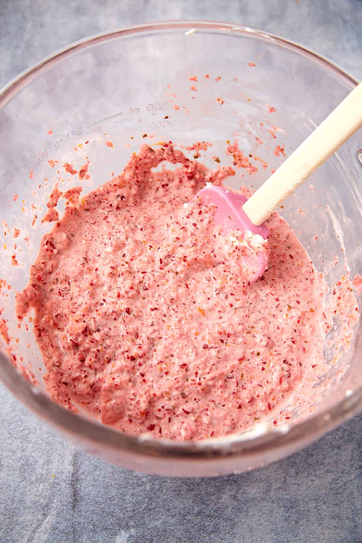 A glass bowl filled with a pink, chunky strawberry mixture. A pink spatula with a wooden handle rests inside the bowl, and the bowl is placed on a gray textured surface.