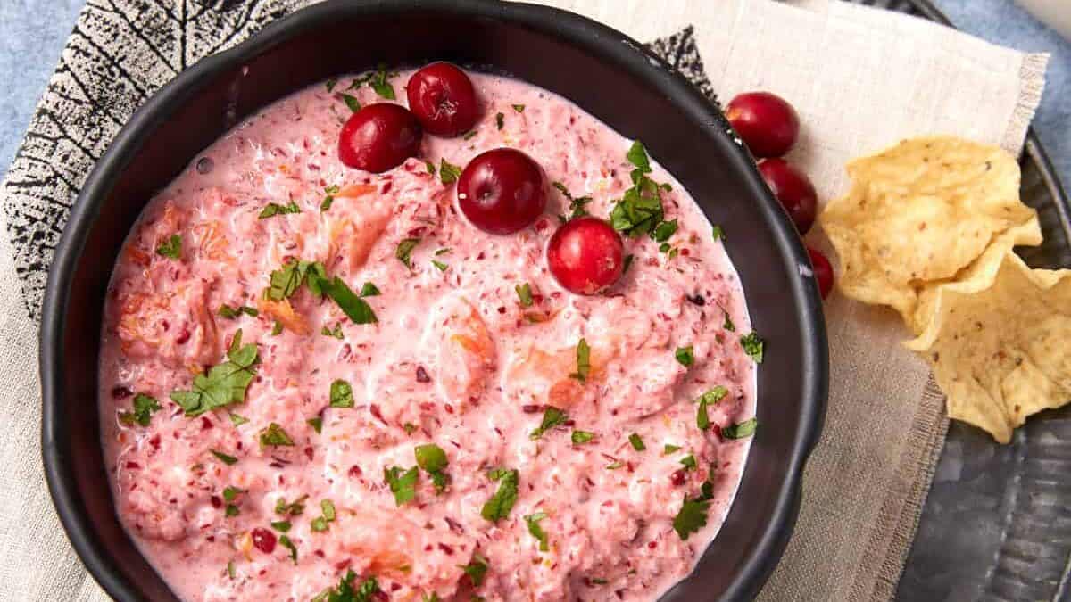A black bowl filled with creamy pink dip garnished with chopped herbs and cranberries, placed on a napkin-covered plate. Tortilla chips and extra cranberries are next to the bowl on a light blue surface.