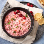 A black bowl filled with creamy cranberry salsa garnished with fresh cranberries and chopped herbs, placed on a beige napkin with tortilla chips on the side.