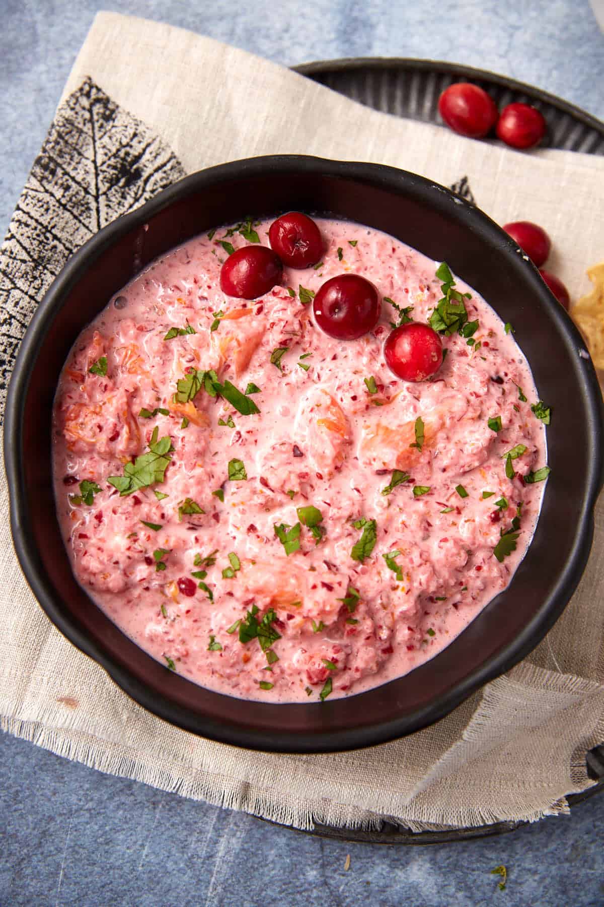 A black bowl filled with a pink cranberry salad, garnished with whole cranberries and chopped herbs, sits on a beige napkin atop a blue surface. A small plate with cranberries is in the background.
