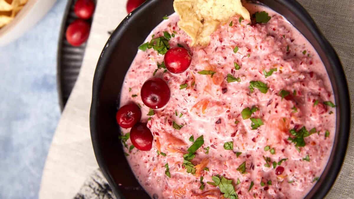 A black bowl filled with creamy cranberry salsa, garnished with whole cranberries and chopped herbs, sits on a light cloth with tortilla chips nearby. A bowl of chips is in the background.