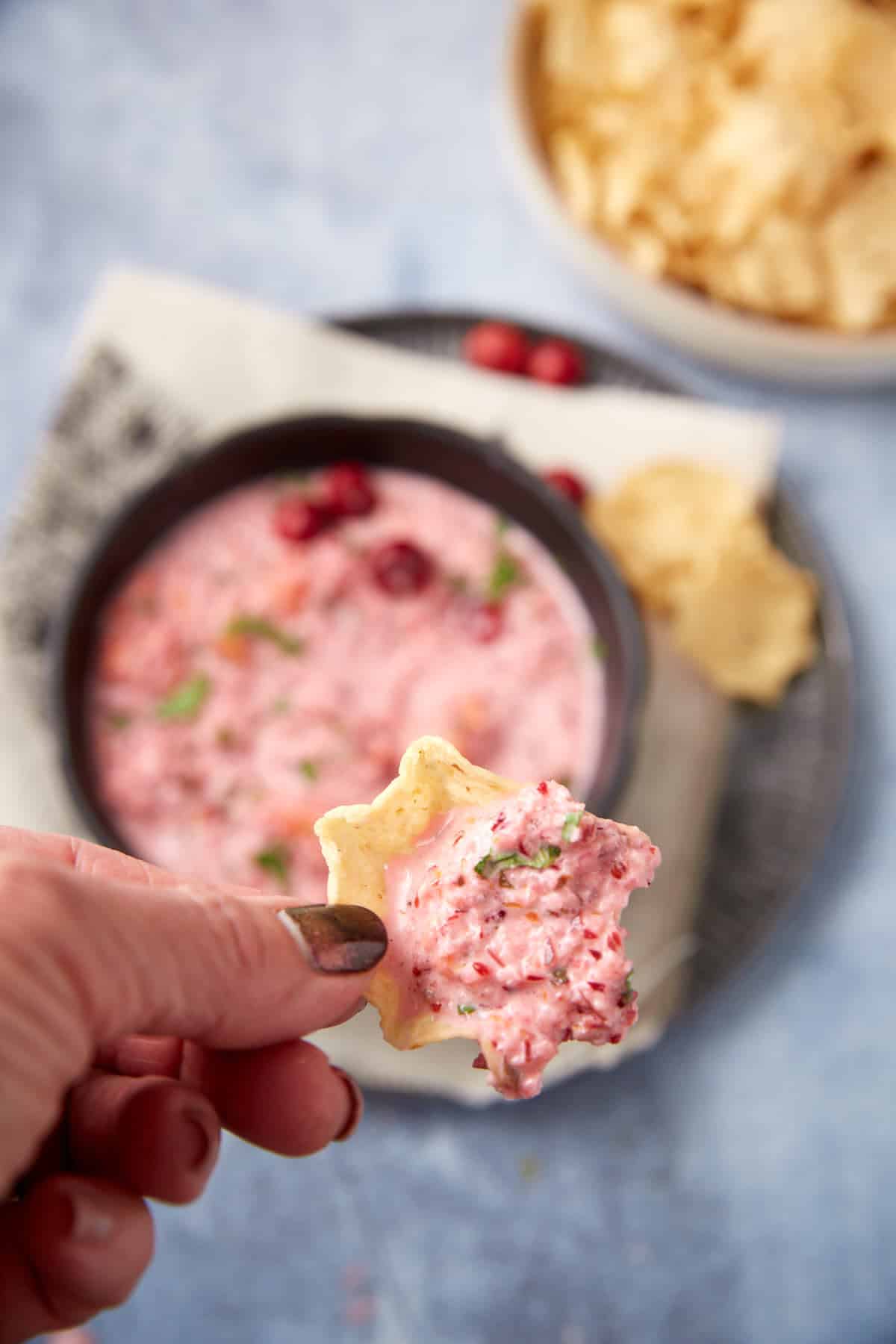 A hand holds a chip topped with creamy pink dip, with a bowl of the same dip and a plate of chips in the background on a light blue surface.